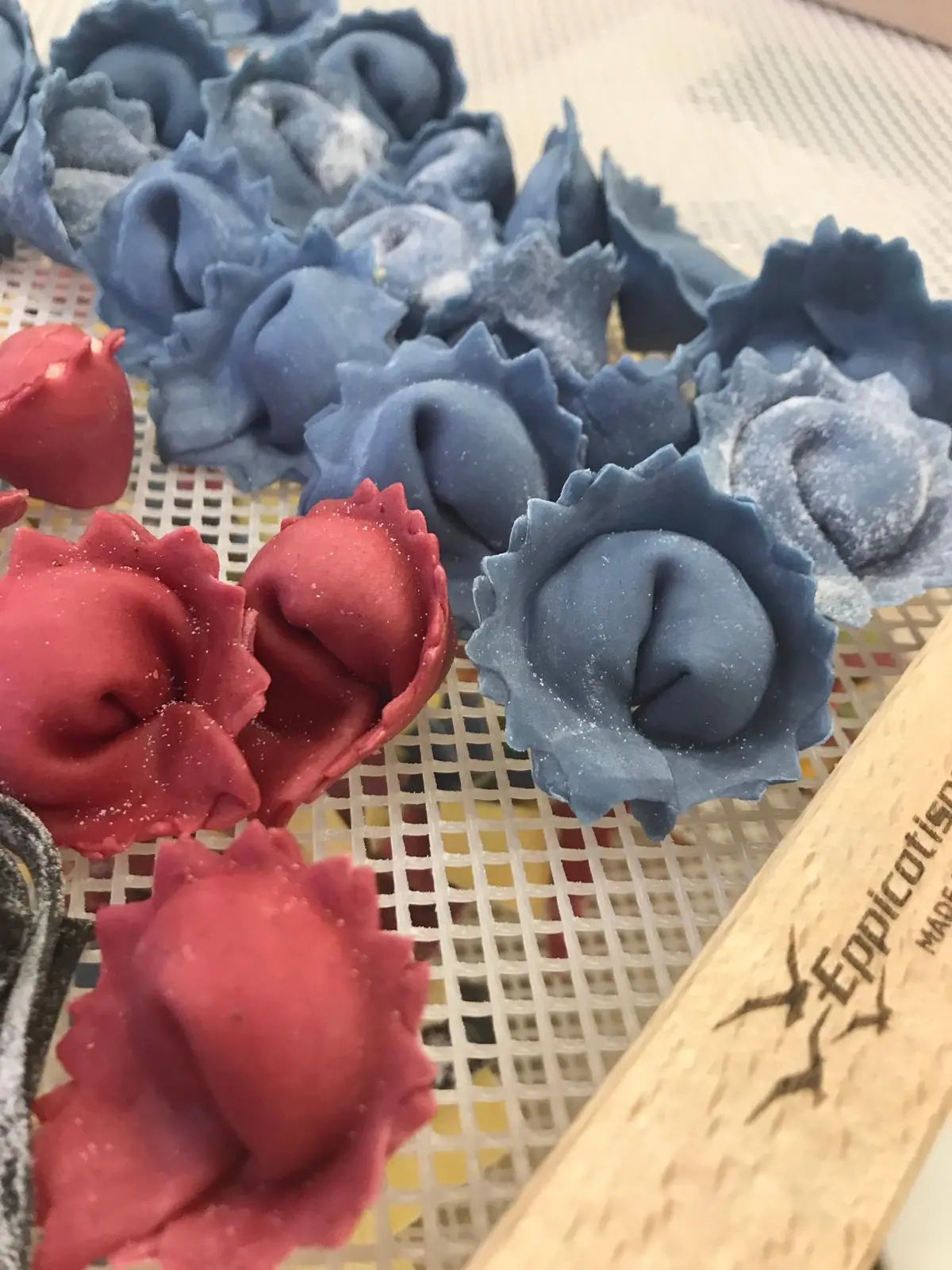 Red and blue rose-shaped ravioli on a textured surface with a wooden block in the foreground.