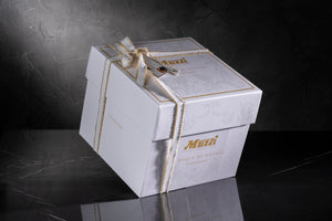 A white rectangular gift box with gold foil detailing and a cream-colored ribbon tied in a bow, featuring the brand name 'Mazi' on the front, set against a dark textured background.