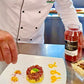 Chef preparing a dish with a bottle of red sauce on a kitchen counter with aleppo chili