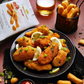 Crispy golden-brown breaded fish sticks served on a black plate with lemon slices and chopped green onions, set on a dark table with a bag of Pizza breadsticks I Bibanesi, fries in a basket, and glasses of beer.