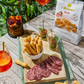 A styled food spread on a wooden table featuring sliced salami, breadsticks, and a bag of i Bibenesi Pizza breadsticks, alongside drinks and greenery in the background.