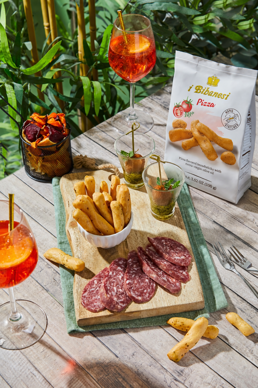 A styled food spread on a wooden table featuring sliced salami, breadsticks, and a bag of i Bibenesi Pizza breadsticks, alongside drinks and greenery in the background.