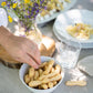 A white bowl filled with small breadsticks I Bibanesi on a table, with a hand reaching in. A glass vase with yellow and purple wildflowers sits behind the bowl, along with plates of food and a clear drinking glass.