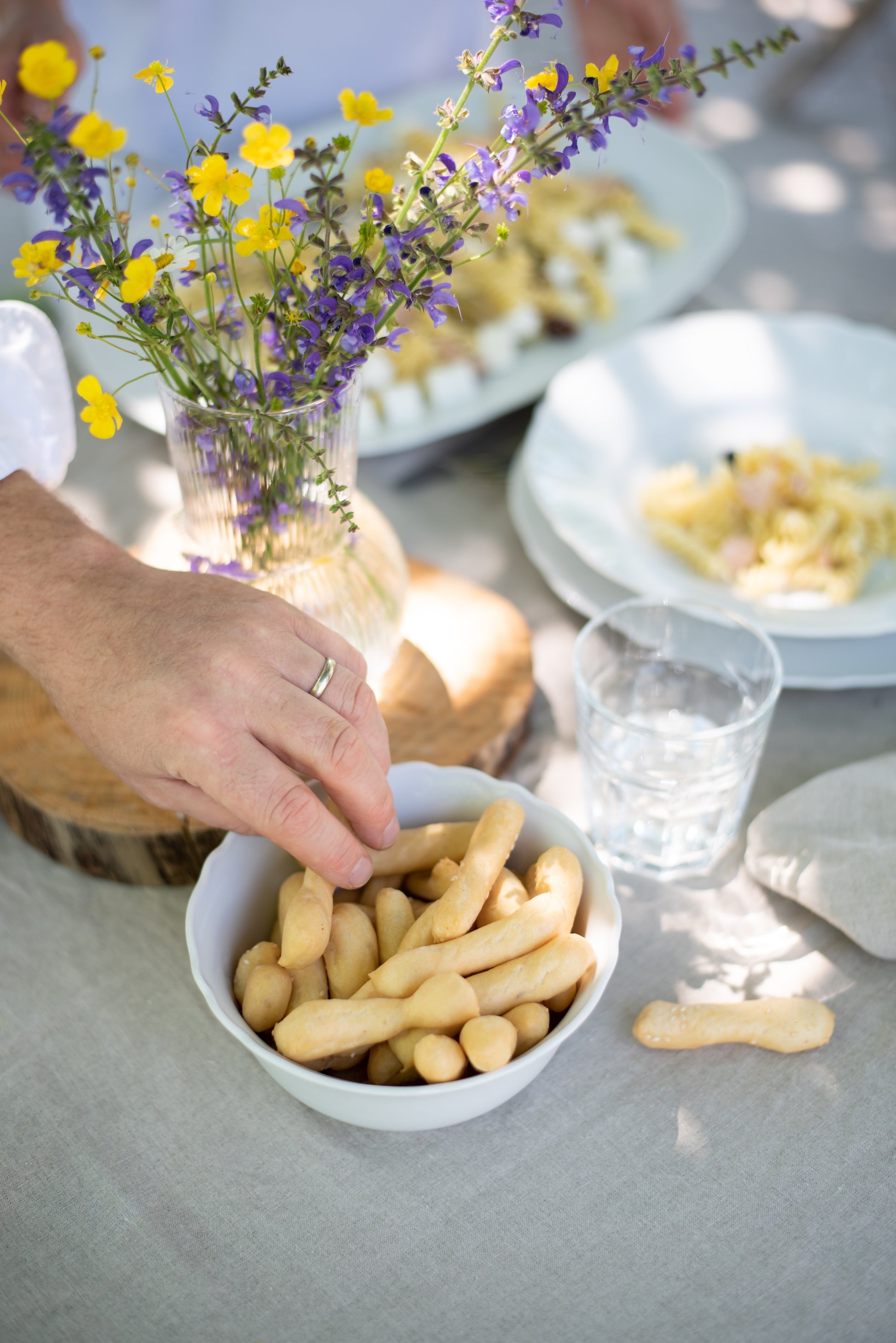 A white bowl filled with small breadsticks I Bibanesi on a table, with a hand reaching in. A glass vase with yellow and purple wildflowers sits behind the bowl, along with plates of food and a clear drinking glass.