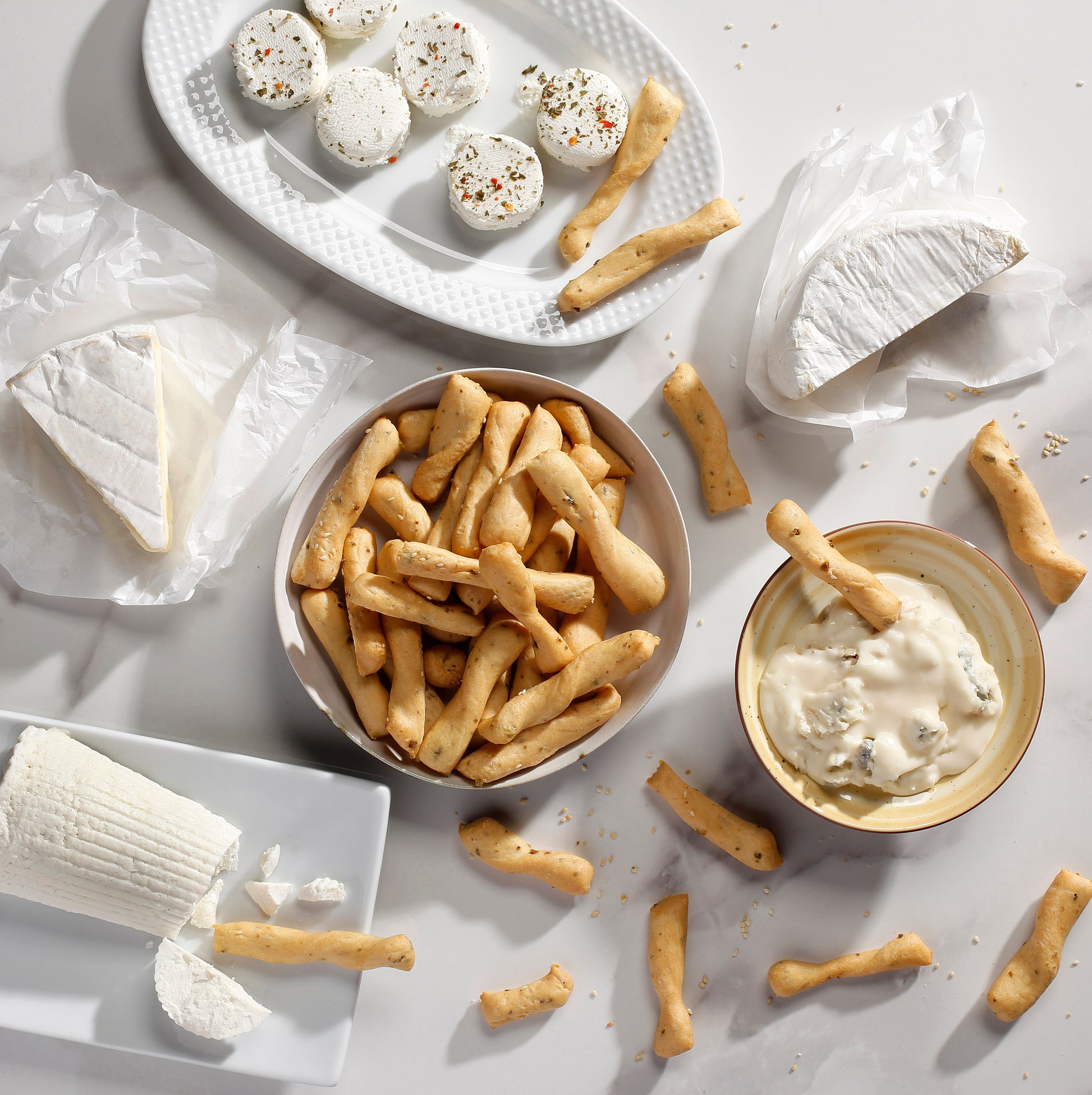 A styled flat-lay of cheese and breadsticks i Bibanesi on a white surface. A bowl holds golden-brown breadsticks, a small dish has creamy white dip with a breadstick, a plate holds round cheese balls with herbs, and other cheese wedges and slices are wrapped in plastic or arranged on a white plate.