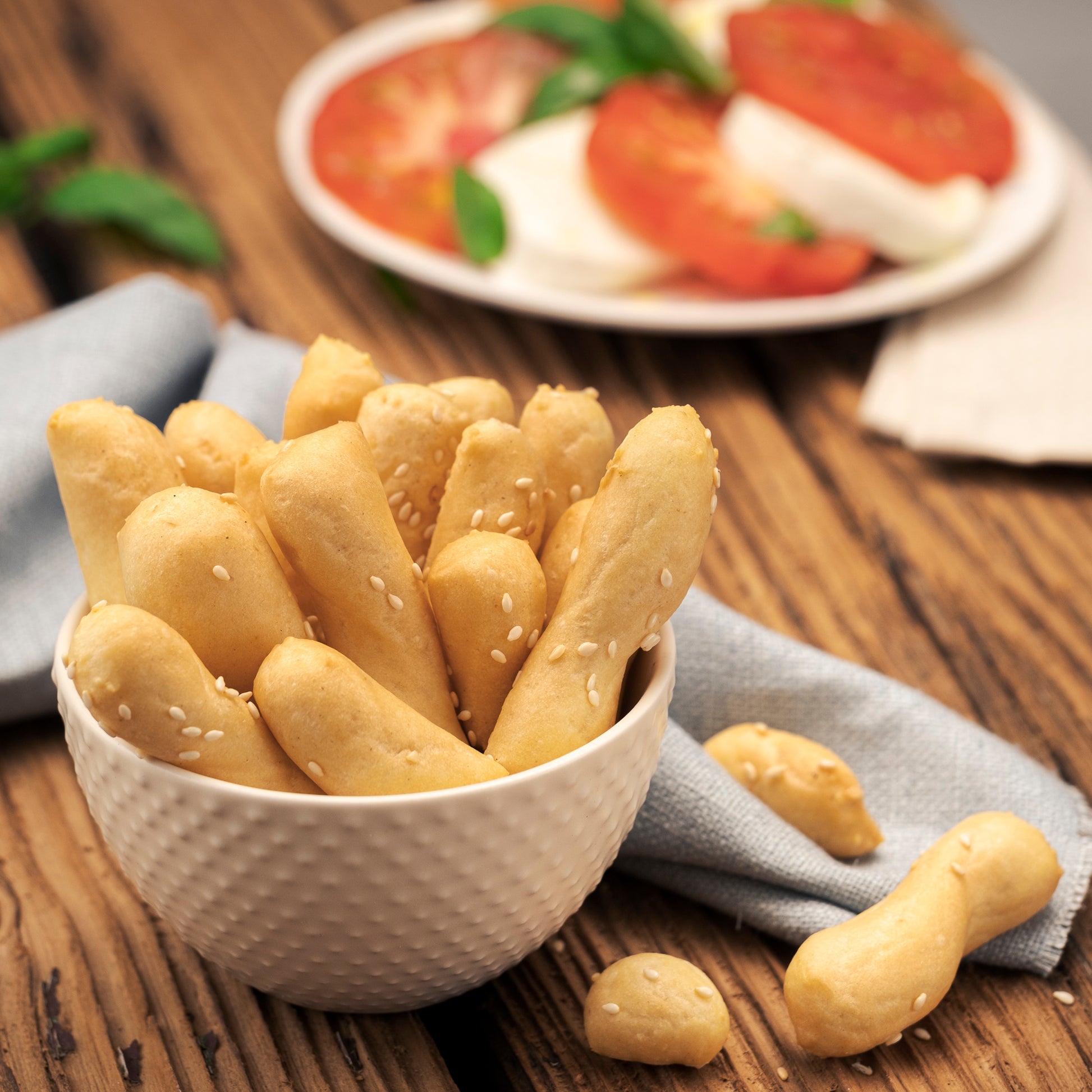 Golden-brown breadsticks i Bibanesi sprinkled with sesame seeds, presented in a textured cream-colored bowl on a wooden table, with a blurred plate of tomato and mozzarella slices in the background.