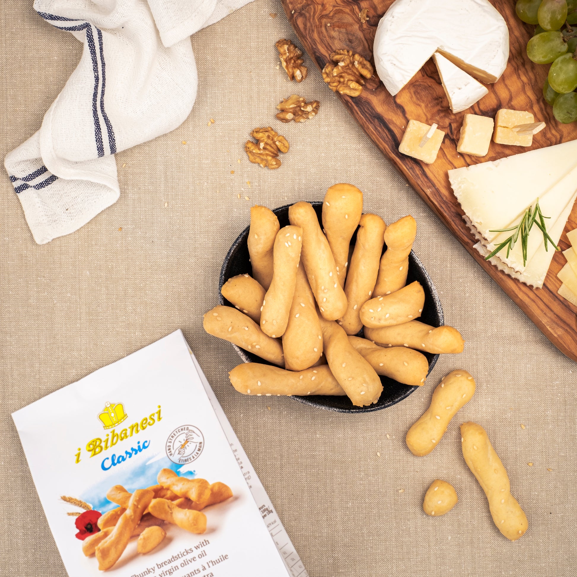 Table with a I bibanesi pack and some breadsticks in a bowl on the table, with a cutting board with some cheeses and green grapes