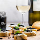 Cured truffle pearls in a glass jar labeled 'PERLÉ DI TARTUFO Truffle Oil' styled on a wooden board with crostini topped with caviar, a glass of white wine, and a bottle in the background.