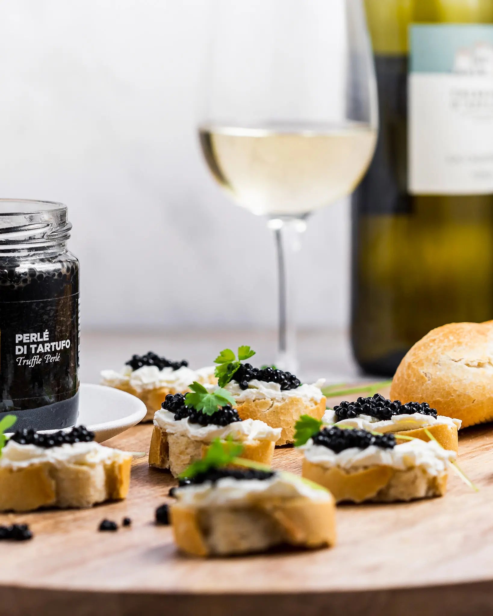 Cured truffle pearls in a glass jar labeled 'PERLÉ DI TARTUFO Truffle Oil' styled on a wooden board with crostini topped with caviar, a glass of white wine, and a bottle in the background.