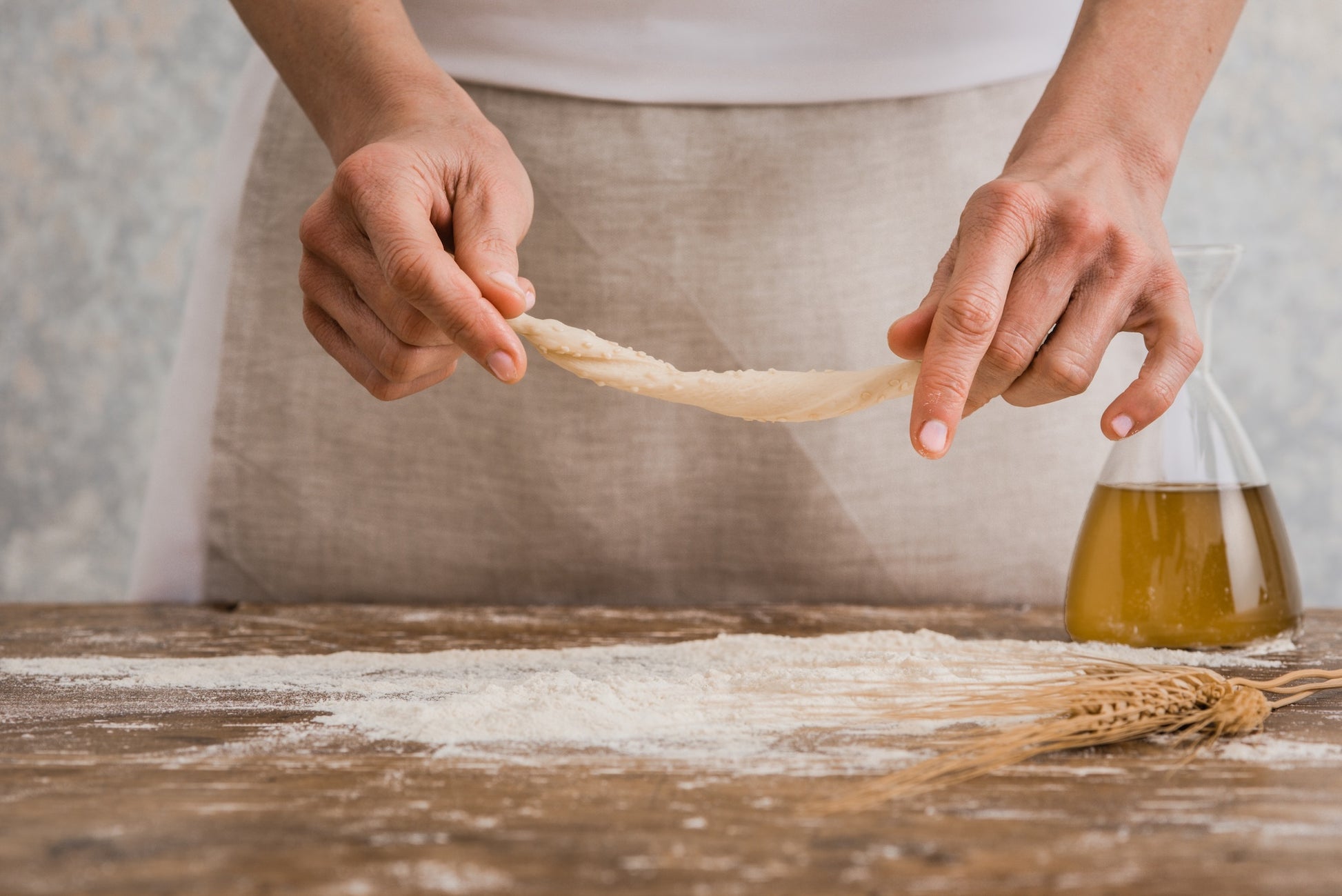 A person's hands stretching a piece of I Bibanesi dough over a rustic wooden table dusted with flour, with a glass beaker of oil and wheat stalks in the background.