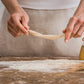 A person's hands stretching a piece of I Bibanesi dough over a rustic wooden table dusted with flour, with a glass beaker of oil and wheat stalks in the background.