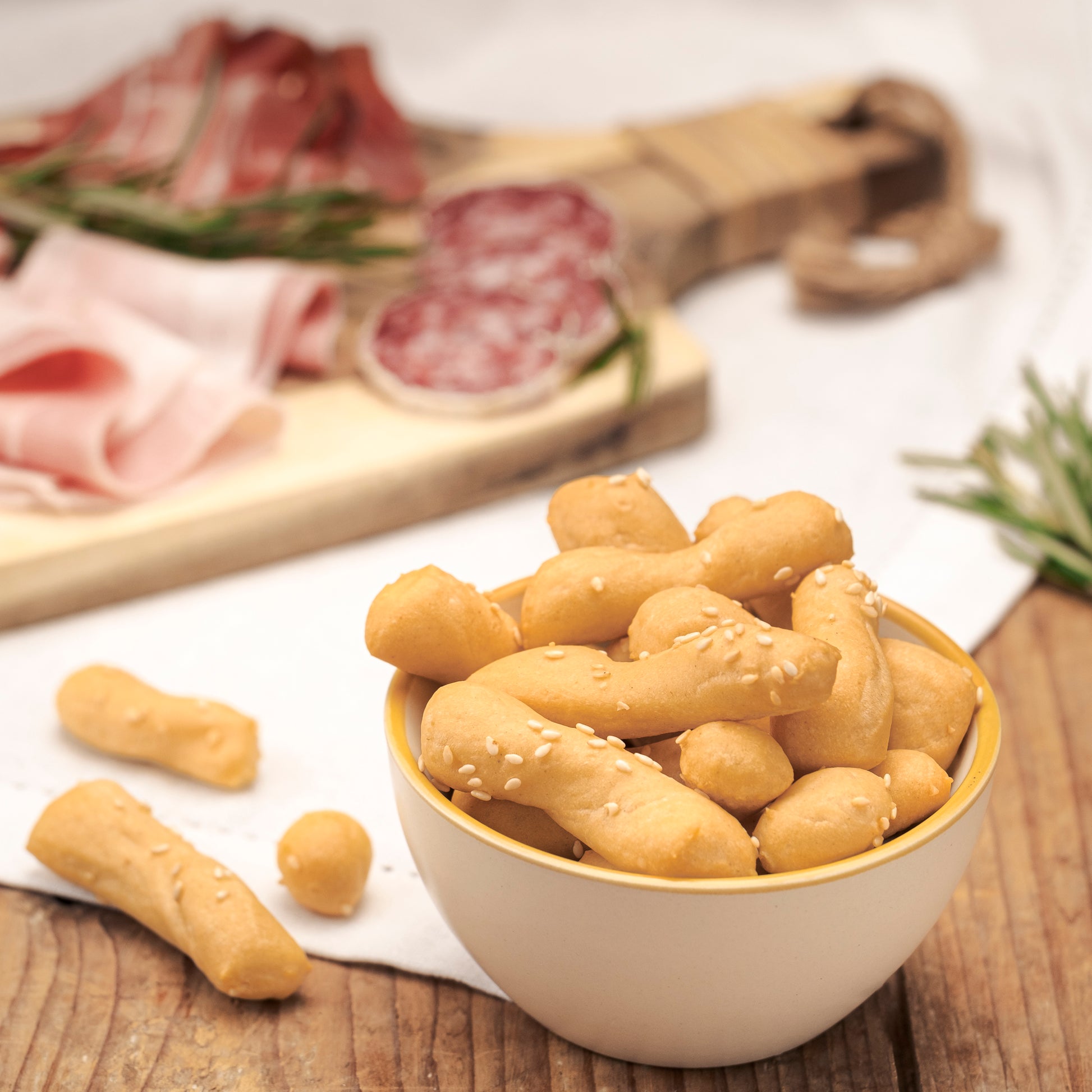 A bowl of golden-brown breadsticks I Bibanesi sprinkled with sesame seeds, set on a wooden table with sliced cured meats and rosemary in the background.