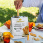 A person's hand reaches into a bag of i Bibanesi Italian Mini Chilli Breadsticks on an outdoor picnic table. The scene features a white tablecloth, plates of food, orange drinks with orange slices, and a blurred green grass background.