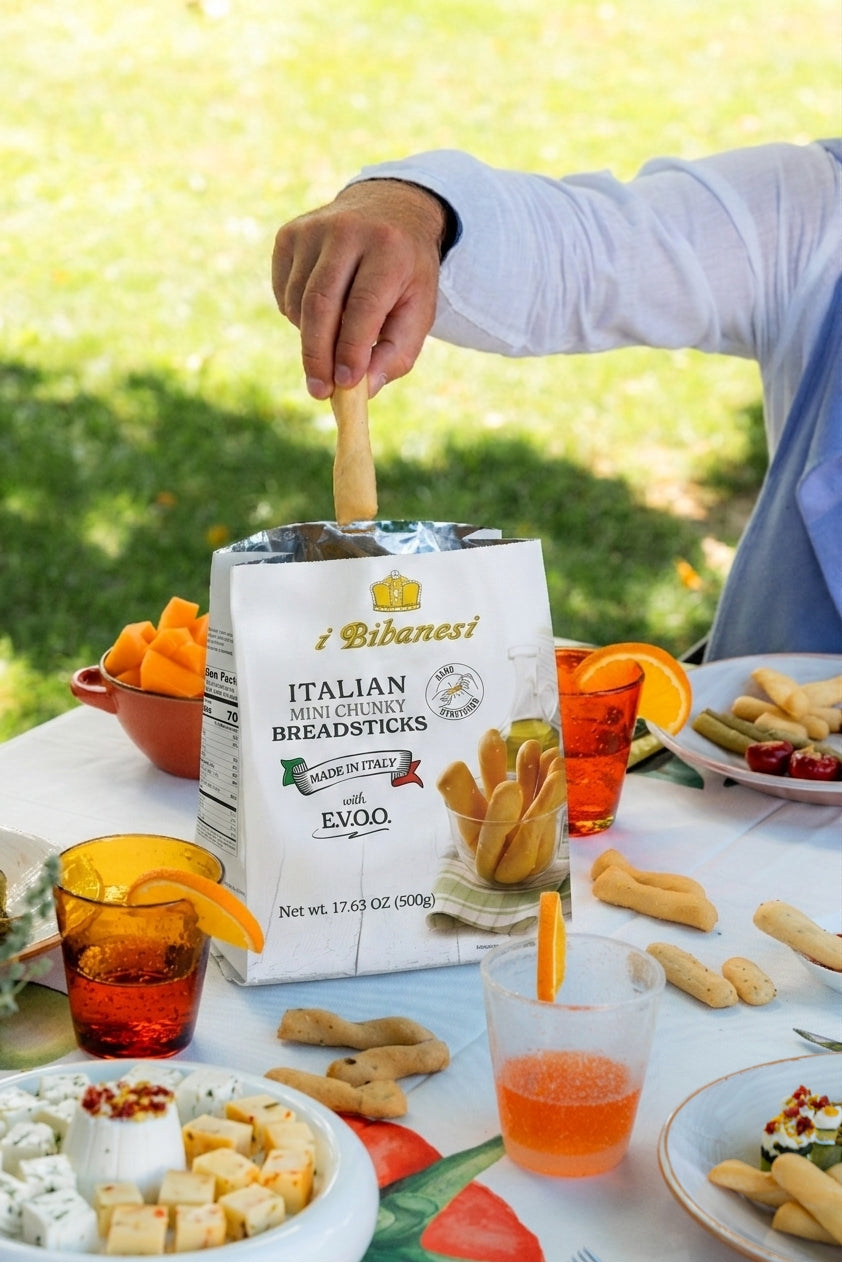 A person's hand reaches into a bag of i Bibanesi Italian Mini Chilli Breadsticks on an outdoor picnic table. The scene features a white tablecloth, plates of food, orange drinks with orange slices, and a blurred green grass background.