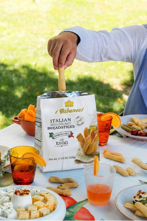 A person's hand reaches into a bag of i Bibanesi Italian Mini Chilli Breadsticks on an outdoor picnic table. The scene features a white tablecloth, plates of food, orange drinks with orange slices, and a blurred green grass background.