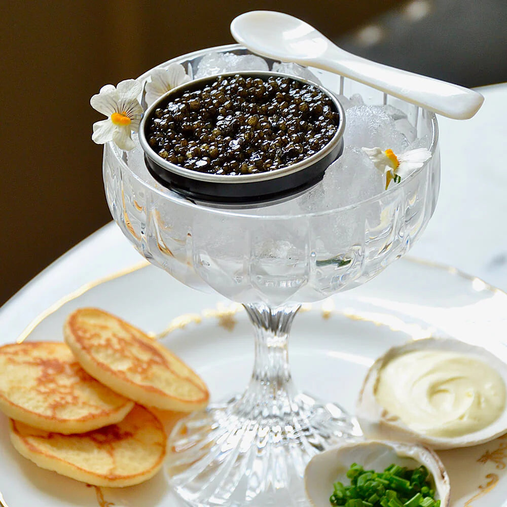 Caviar in a decorative glass dish with crackers and condiments on a white plate.