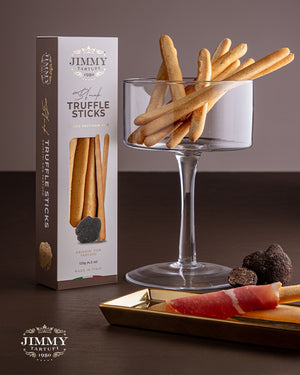 breadsticks artisanal truffle crackers shown with their branded box, served in a clear glass platter, and paired with cured ham and truffles on a gold tray against a dark brown background.