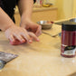 Person kneading dough on a kitchen counter with a jar of red food coloring nearby.