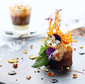 Delicate savory cake with floral garnishes on a gray plate, accompanied by a glass of iced coffee.