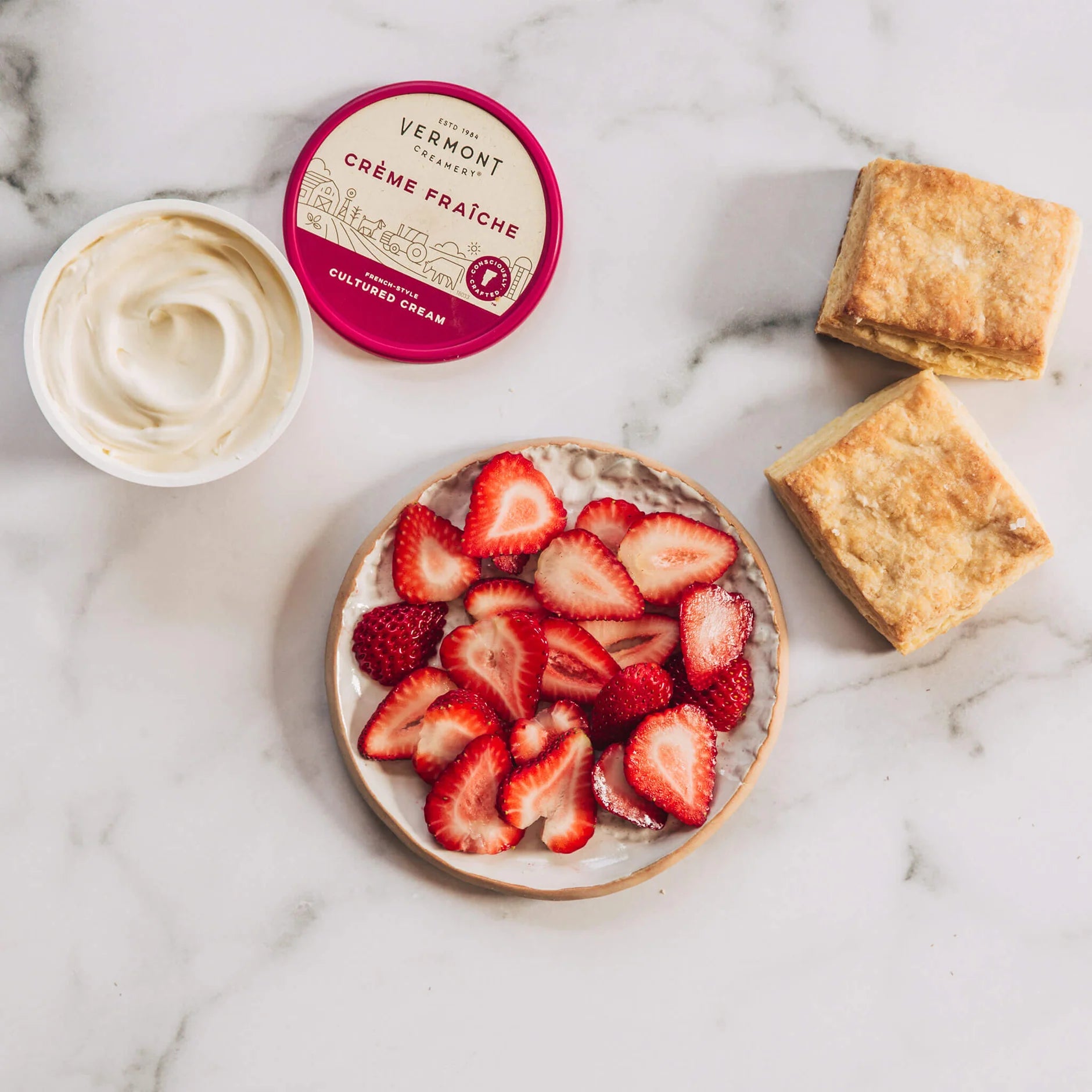 Bowl of strawberries, container of Vermont Crème Fraçè, and biscuits on a marble surface
